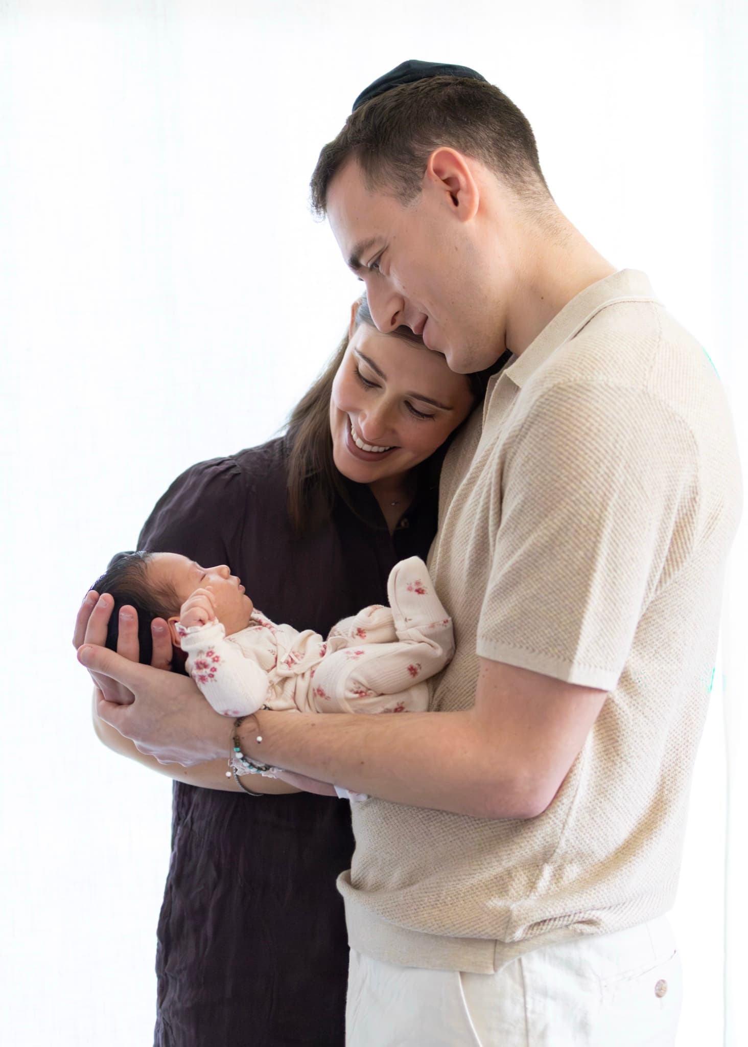 Parents cradling their newborn baby in a floral romper by the window — natural light newborn photography