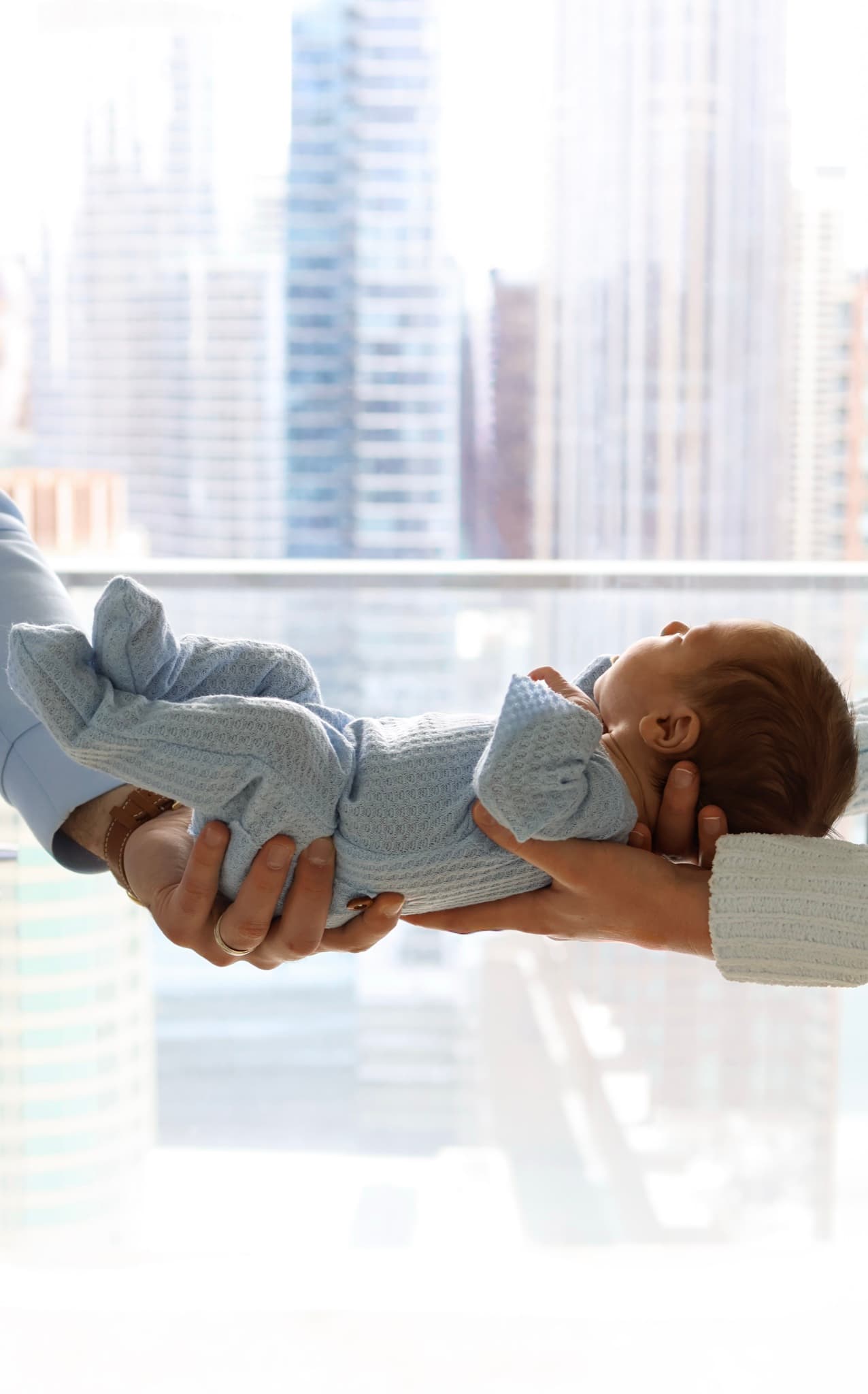 in-home newborn photographer Skokie — parents' hands cradling sleeping baby by the window with city skyline